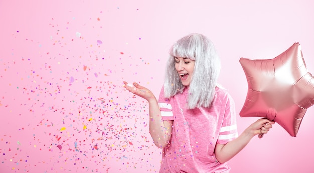 Funny Girl con cabello plateado da una sonrisa y emoción en la pared rosa. Joven mujer o niña adolescente con globos y confeti