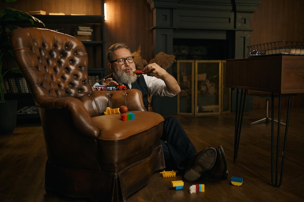 Foto funny hombre de negocios senior jugando juguetes disfrutando de un tiempo divertido después de un duro día de trabajo en la oficina de casa. hombre mayor sentado en el suelo cerca del sillón
