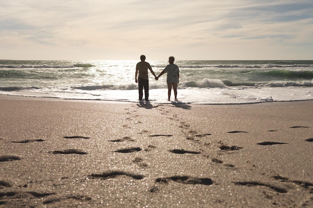 Fußabdrücke im Sand mit einem gemischtrassigen älteren Paar, das an einem sonnigen Tag Händchen haltend am Strand steht