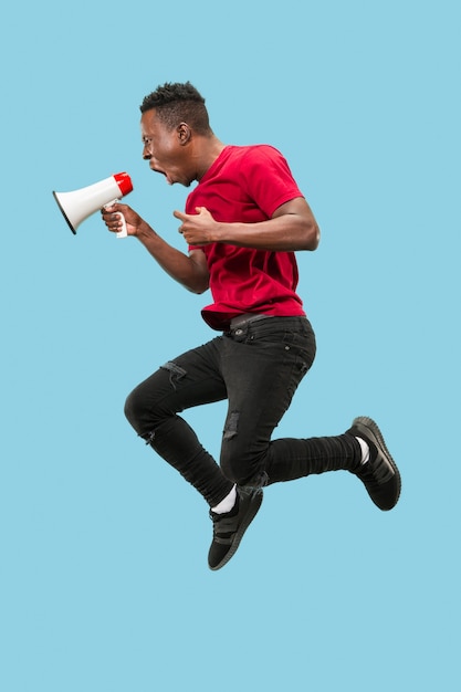 Fußballfan, der auf orangefarbenem Hintergrund springt. Der junge afrikanische Mann als Fußballfan mit Megaphon isoliert auf blauem Studio. Unterstützungskonzept. Menschliche Emotionen, Gesichtsausdruckkonzepte.