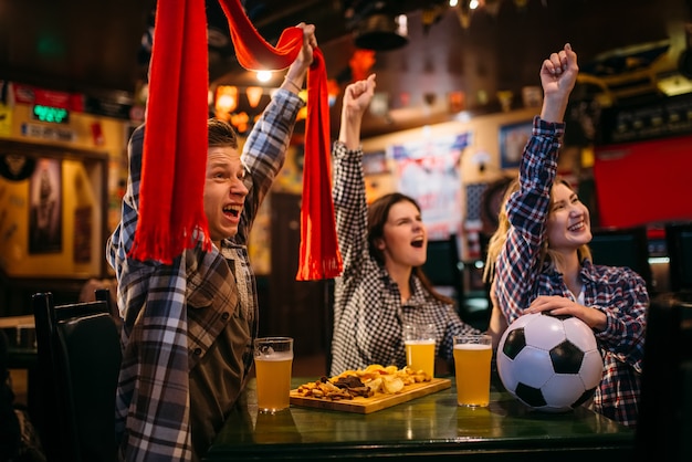 Fußballfans mit Schal schauen sich das Spiel an und heben die Hände in der Sportbar. Fernsehsendung, Freizeitfreunde junger Freunde in der Kneipe, Lieblingsteam gewinnt