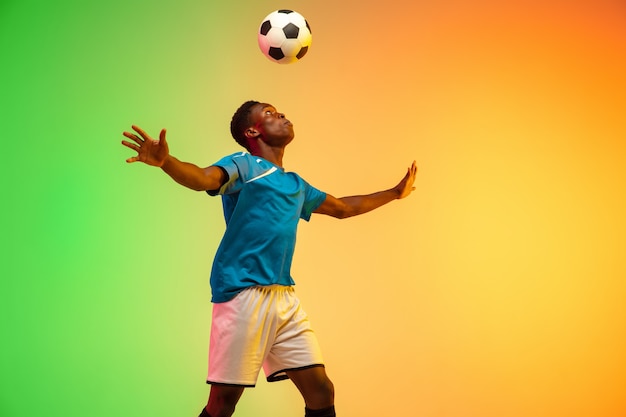 Fútbol masculino afroamericano, formación de jugador de fútbol en acción aislada sobre fondo de estudio degradado en luz de neón. Concepto de movimiento, acción, logros, estilo de vida saludable. Cultura juvenil.