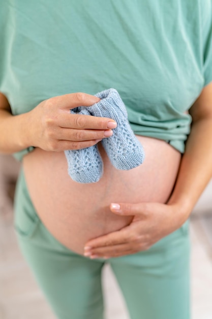 La futura mamá con ropa informal se pone de pie y sostiene calcetines para el recién nacido en su vientre. Su mano izquierda cubre el ombligo. Vista desde arriba.