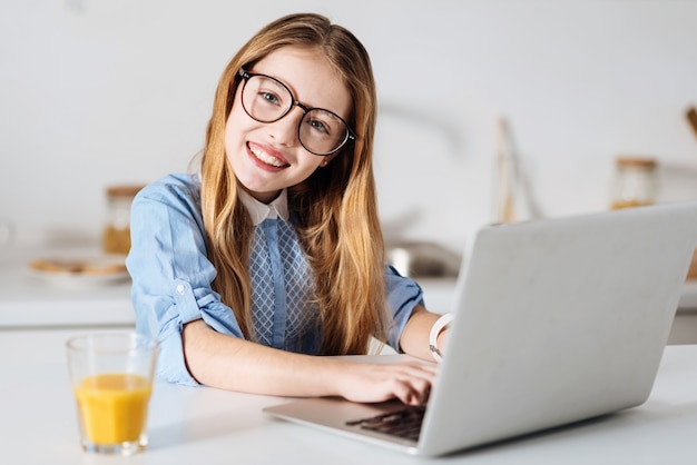 Futuro autor. Encantadora niña activa optimista escribiendo un ensayo usando su computadora mientras trabajaba en la tarea del hogar