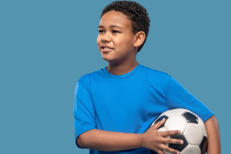 Futuro jogador de futebol. Menino alegre e interessado, de pele escura, usando uma camisa esportiva azul, segurando uma bola de futebol em pé no estúdio