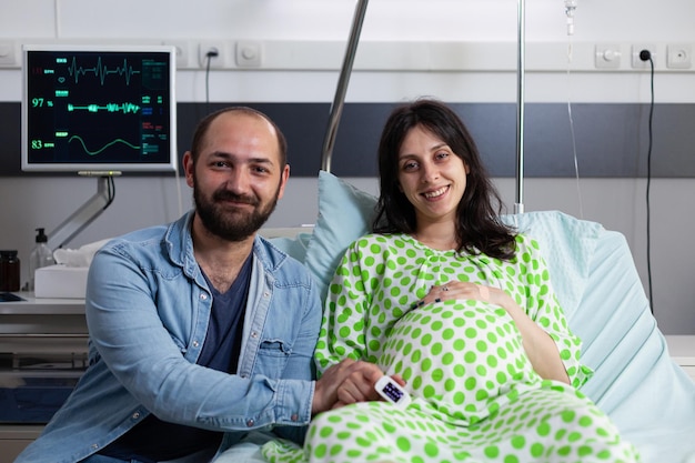 Futuros pais sorridentes esperando criança na enfermaria do hospital, preparando-se para a cirurgia cesariana. Mulher grávida olhando para a câmera enquanto estava deitado na cama esperando o médico na maternidade. conceito de parto