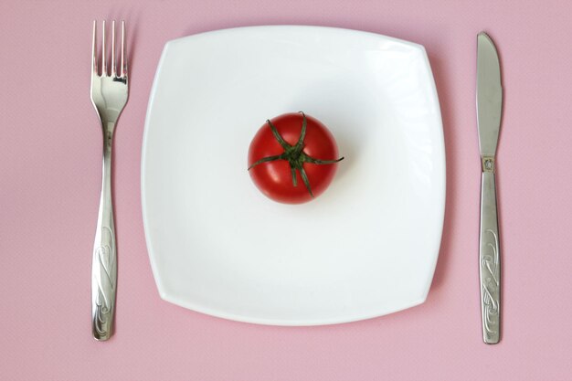 Gabelmesser Tomate und weißer Teller auf rosa Hintergrund Sauberer Teller und Besteck auf rosa Hintergrund