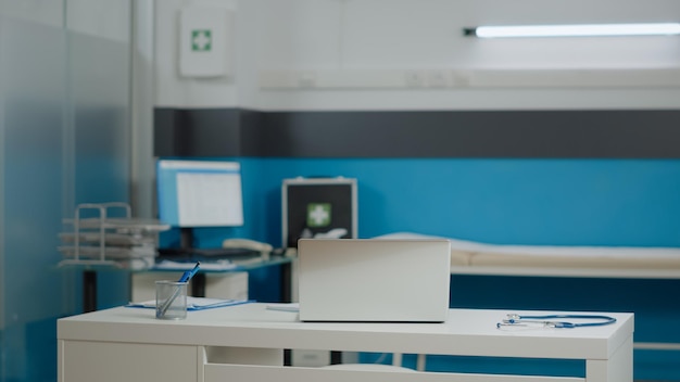 Gabinete médico vacío en el centro de salud con laptop, estetoscopio y papeles de chequeo en el escritorio blanco. Nadie en el interior profesional para consultar con muebles y herramientas modernas.