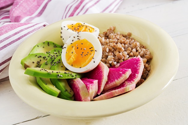 Gachas de alforfón Buda cuencos con aguacate, huevos duros y rábano de sandía en mesa blanca. Desayuno saludable.