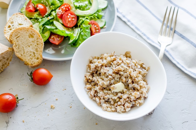 Gachas de alforfón caliente con mantequilla derretida en un plato blanco y ensalada de verduras para lanzar en la mesa, vista superior, espacio de copia Concepto de comida sana