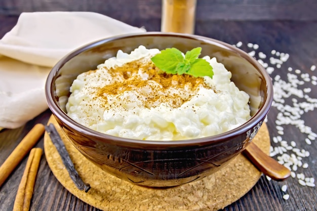 Gachas de arroz con canela, menta en un recipiente marrón, servilleta, cuchara, vaina de vainilla sobre una tabla de madera oscura.