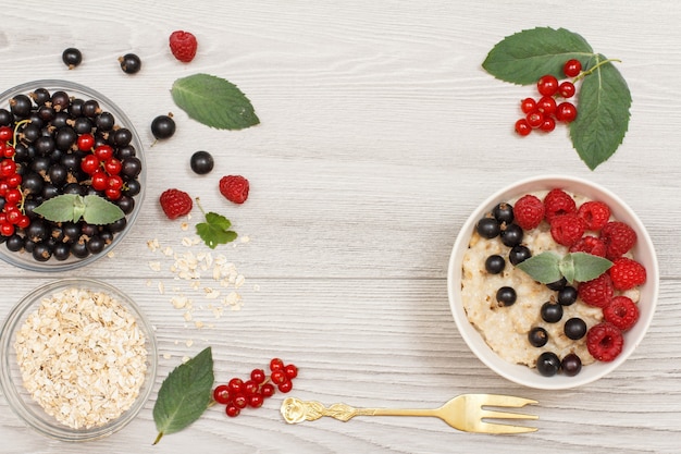 Gachas de avena en cuenco de porcelana con grosellas y frambuesas, decoradas con hojas de menta. Cuencos de vidrio con copos de avena y grosellas con tenedor de metal. Vista superior con espacio de copia.