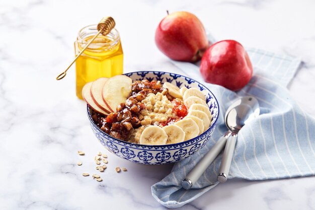 Gachas de avena con manzanas caramelizadas con canela, plátano, fresas ralladas y miel sobre fondo de mármol claro