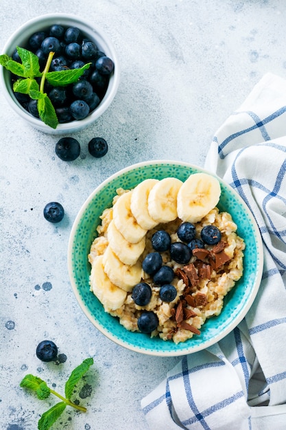 Gachas de avena con plátano, chocolate y arándanos frescos en un recipiente sobre una superficie de pizarra, piedra u hormigón gris claro