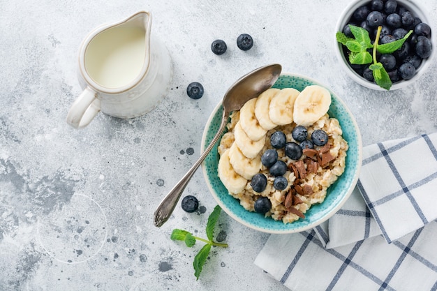 Gachas de avena con plátano, chocolate y arándanos frescos en un recipiente sobre una superficie de pizarra, piedra u hormigón gris claro