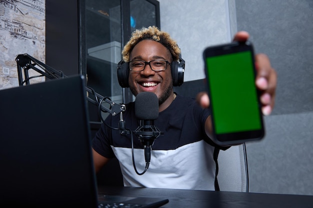 Gadget com espaço livre vazio para um layout um banner Retrato de um negro sorridente sentado em frente ao microfone e usando fones de ouvido segurando na mão um grande smartphone com tela verde em branco