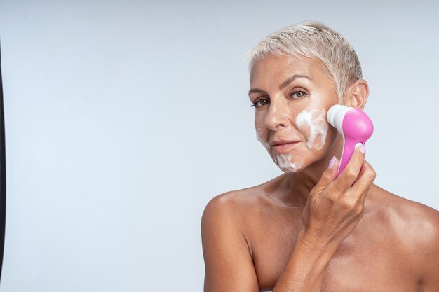 Gadget moderno. Mujer alegre limpiando su rostro, cuidando su piel envejecida