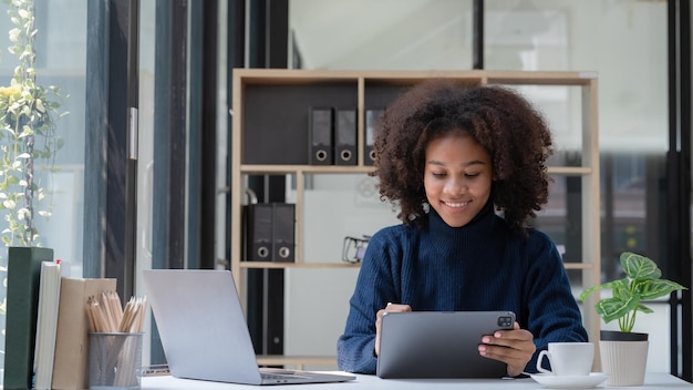 Gadgets de negocios Mujer de negocios negra feliz usando una tableta digital y una computadora portátil trabajando en una oficina moderna