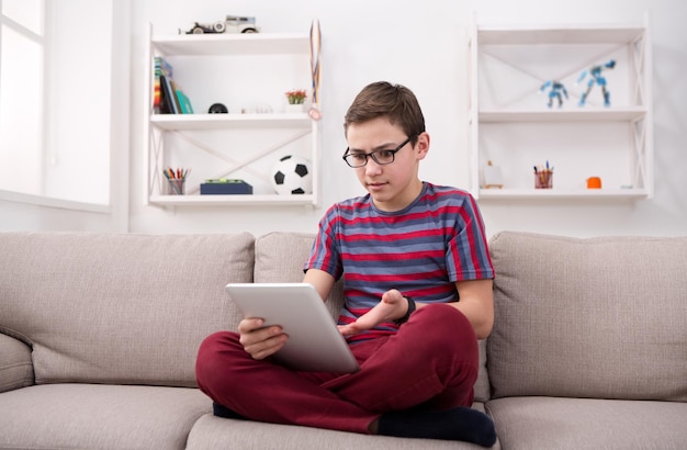 Gadgets und Technologiekonzept - Teenager mit digitalem Tablet, Fußball auf tragbarem Gerät gucken, Online-Spiel spielen, während er zu Hause auf der Couch im Wohnzimmer sitzt