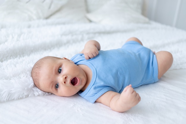Gähnender kleiner Junge drei Monate alt in einem blauen Body auf einem weißen Bett zu Hause geht ins Bett