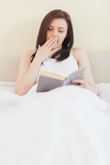 Gähnendes Mädchen, das ein Buch liegt auf einem Bett liest