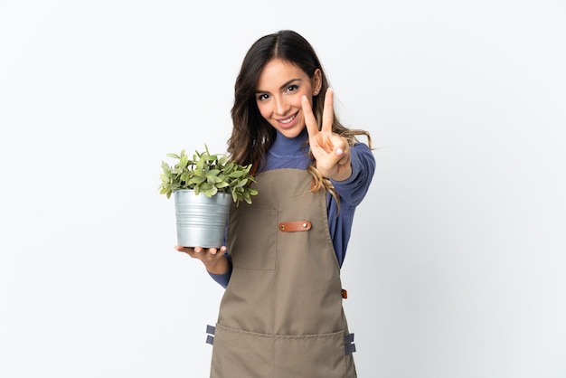Gärtnermädchen hält eine Pflanze lokalisiert auf weißem Hintergrund lächelnd und zeigt Siegeszeichen