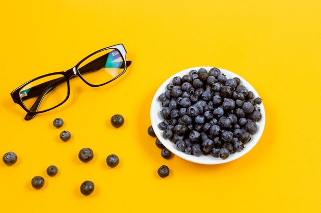 Gafas y arándanos frescos en plato blanco sobre fondo amarillo