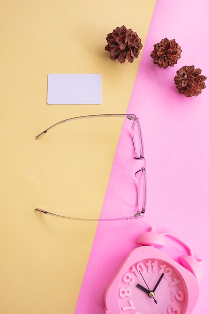 Gafas cuadradas en la foto de estilo minimalista veraniego sobre un fondo rosa pastel y amarillo. Reloj despertador, flores de pino, tarjetas de visita