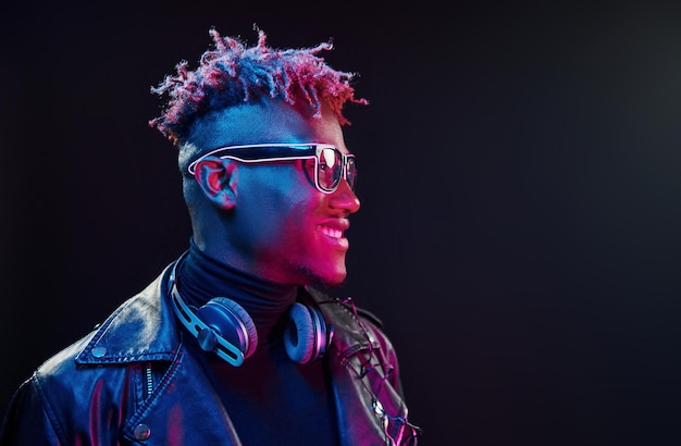 Gafas con estilo y auriculares inalámbricos Iluminación de neón futurista Joven afroamericano en el estudio