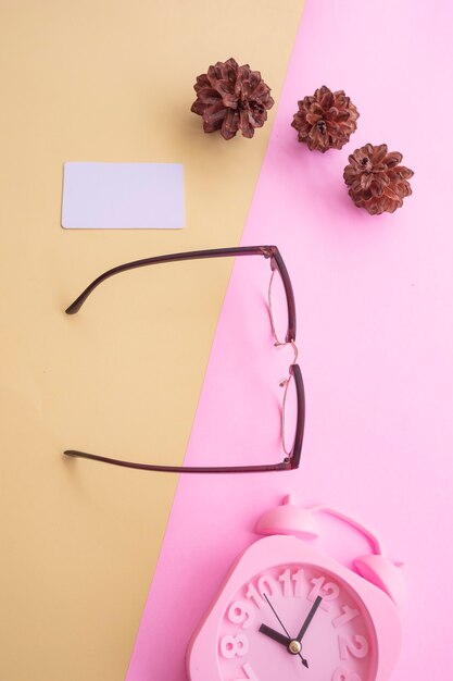 Gafas con estilo sobre fondo pastel. Tarjeta de visita, colores pastel rosa y amarillo. Reloj despertador, flores de abeto.