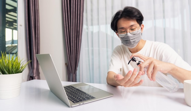 Gafas de hombre guapo joven de negocios asiático que trabaja desde la oficina en casa pone en cuarentena el coronavirus de la enfermedad o COVID-19 usando máscara protectora y limpiando las manos con gel desinfectante en la computadora portátil delantera