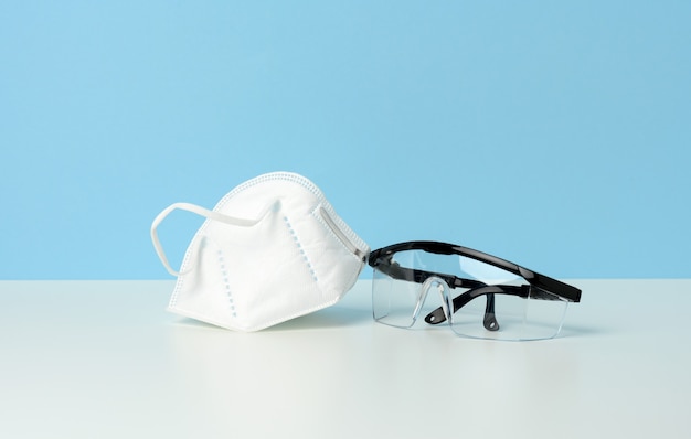 Gafas médicas de protección de plástico transparente y máscara desechable blanca sobre un fondo azul.