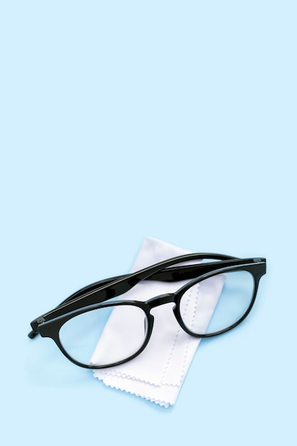 Gafas con montura negra y paño de limpieza sobre fondo azul vertical con espacio de copia. Tejido blando blanco para frotar y pulir las ópticas de la suciedad y el polvo. Corrección de visión, oftalmología, optometría.