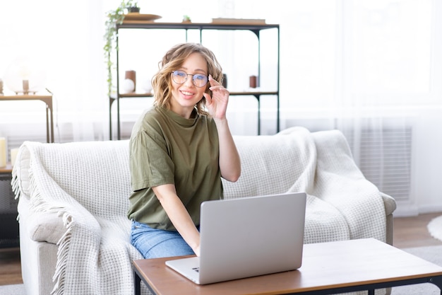 Gafas de mujer con ordenador portátil mientras está sentado en un sofá con ventana grande en el fondo en el interior de una casa