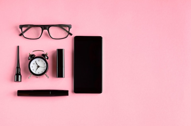 Gafas negras, reloj despertador, teléfono celular, rímel y pomada en superficie rosa.