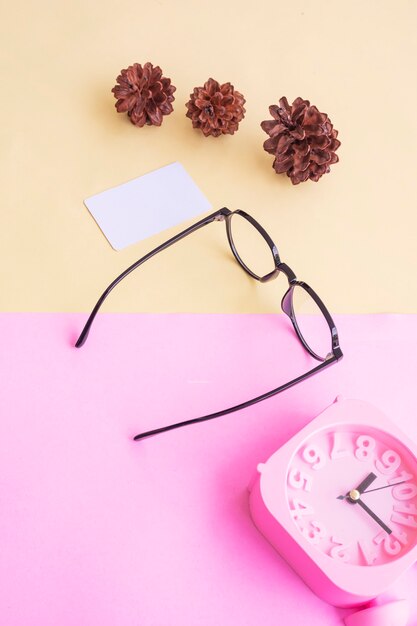 Gafas ovaladas en la foto de estilo minimalista veraniego sobre un fondo rosa pastel y amarillo. Reloj despertador, flores de pino, tarjetas de visita