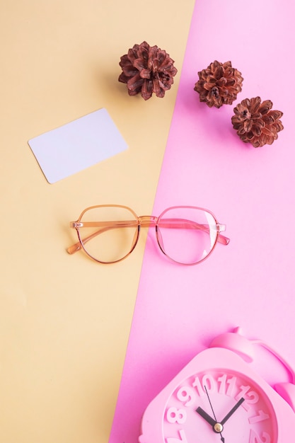 Gafas ovaladas en la foto de estilo minimalista veraniego sobre un fondo rosa pastel y amarillo. Reloj despertador, flores de pino, tarjetas de visita