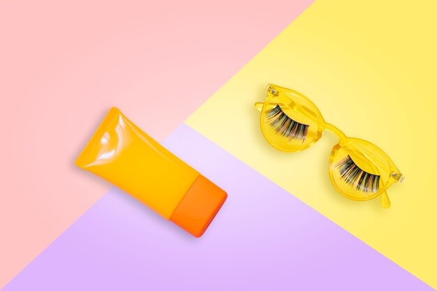 Gafas de sol amarillas con pestañas postizas y crema de protección solar naranja spf sobre fondo rosa.