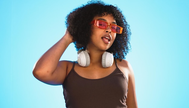 Gafas de sol de guiño facial y cuidado del cabello de la mujer en el estudio aislado en un fondo azul Tonos retro de retrato Gen z y feliz modelo femenino de raza mixta con tratamiento de salón para belleza y auriculares