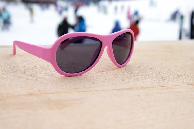 Gafas de sol con montura rosa en pendiente de madera en apres ski bar o cafetería, con pista de esquí en el fondo, copie el espacio. Concepto de deportes de invierno, ocio, recreación, relajación.