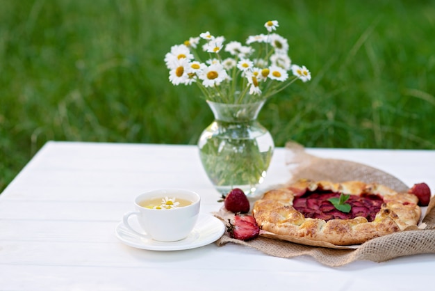 Galette caseiro recém-assado ou torta de morango aberta, uma xícara de chá de ervas e um vaso com um buquê de flores da margarida.