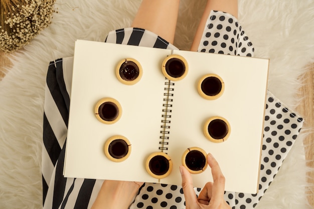 Foto galleta de chocolate en la mano de una dama con lunares y vestido de rayas sobre un libro vacío con galletas de chocolate colocadas en forma de círculo sentado sobre una alfombra blanca de piel esponjosa