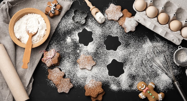 Galleta de jengibre horneada en forma de estrella espolvoreada con azúcar en polvo sobre una mesa negra e ingredientes, vista superior