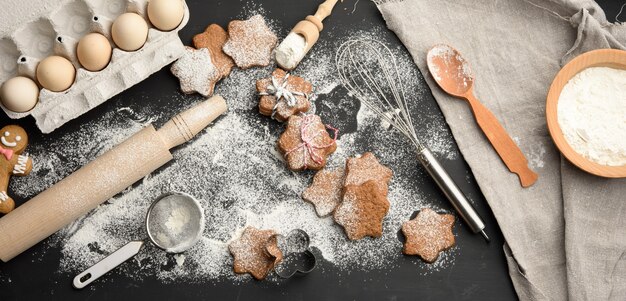 Galleta de jengibre horneada en forma de estrella espolvoreada con azúcar en polvo sobre una mesa negra e ingredientes, vista superior