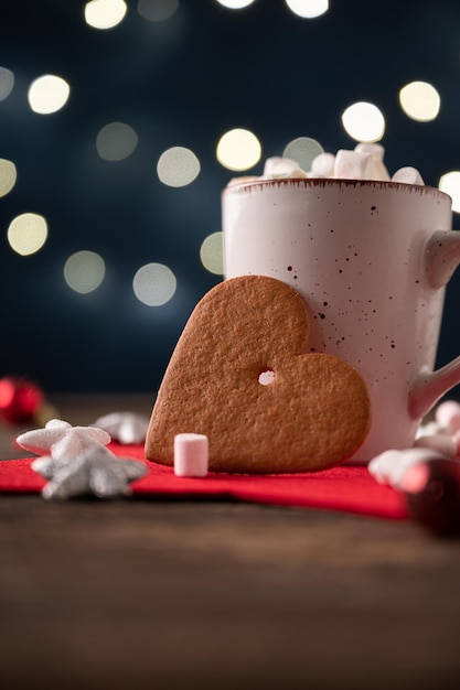 Galleta navideña en forma de corazón grande apoyado en una taza de chocolate caliente con malvaviscos en un entorno navideño con luces brillantes.