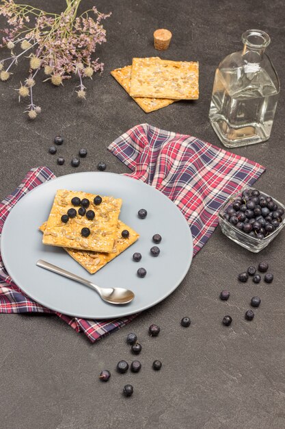 Galletas y arándanos en placa gris sobre servilleta a cuadros. Agua embotellada. Arándanos en un tazón de vidrio. La cuchara está en el plato. Fondo negro. Vista superior