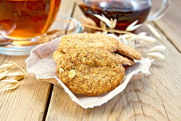 Galletas de avena con un tallo de avena en el papel, una taza de té y una tetera de fondo de tablas de madera