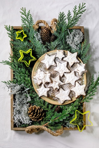 Galletas caseras de cacao y almendras en forma de estrella con glaseado blanco y azúcar glass. En placa de cerámica con cortadores de galletas de estrellas de Navidad, ramas de thuja, decoraciones sobre mantel blanco. Endecha plana