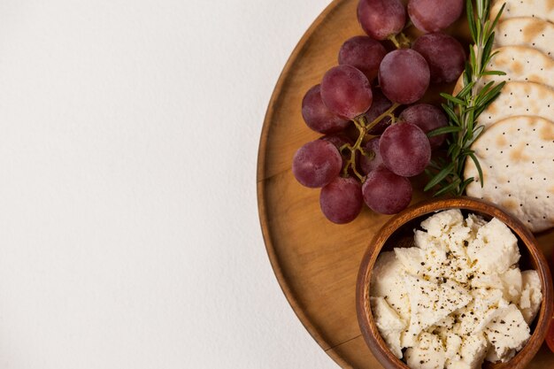 Galletas crujientes, tomates cherry, uvas y tazón de queso sobre tabla de madera