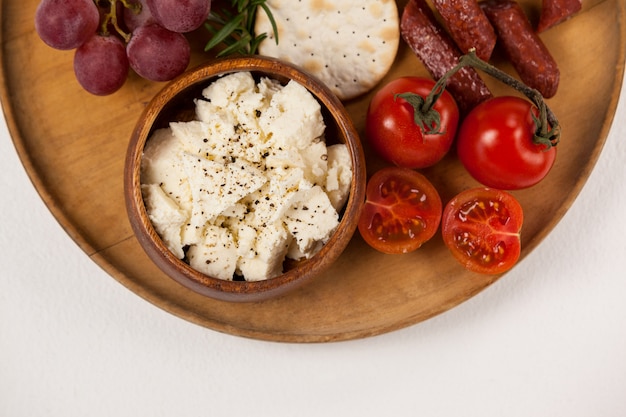 Galletas crujientes, tomates cherry, uvas y tazón de queso sobre tabla de madera